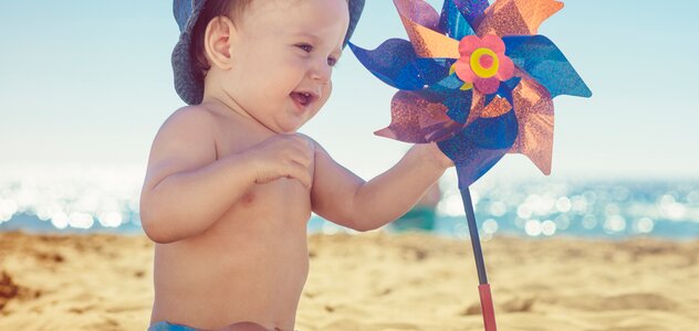 Kilku miesięczny bobas samodzielnie siedzi na plaży. Dziecko ubrane w pieluszkę i kapelusik siedzi na piasku w cieniu parasola. Niemowlę bawi się wiatrakiem, który wbity jest w ziemię.