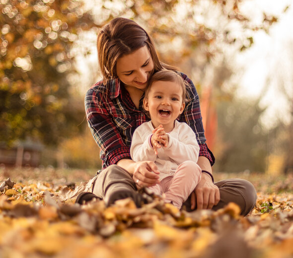 Młoda mama przebywa wraz ze swoim kilkunastomiesięcznym bobasem w parku. Rodzicielka siedzi pośród żółtych i czerwonych liści. Na kolanach kobiety siedzi rozradowana dziewczynka.