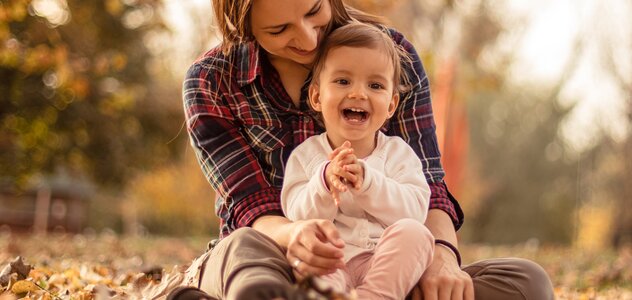 Młoda mama przebywa wraz ze swoim kilkunastomiesięcznym bobasem w parku. Rodzicielka siedzi pośród żółtych i czerwonych liści. Na kolanach kobiety siedzi rozradowana dziewczynka.