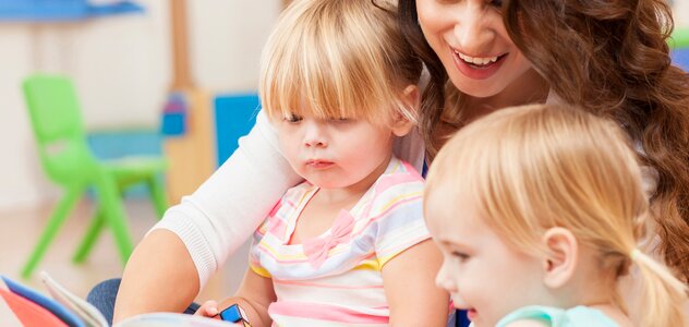 Młoda mama przebywa w pokoju zabaw wraz ze swoimi dwiema młodymi córeczkami. Kobieta siedzi na podłodze i trzyma na kolanach jedną z dziewczynek. Obok rodzicielki siedzi starze dziecko i czytają książkę.