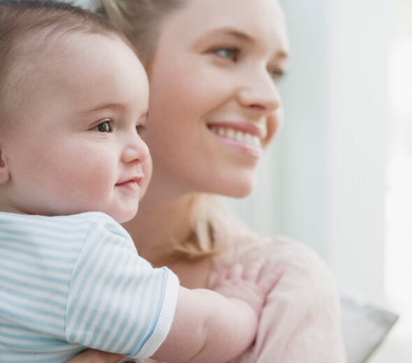 Szczęśliwa młoda mama stoi i trzyma na rękach niedawno narodzone niemowlę. Rodzicielka i bobas patrzą w tę samą stronę wyglądając za okno. Dziecko opiera się rączkami o barki mamy.