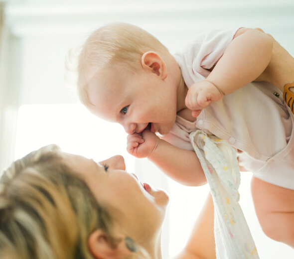 Młoda mama spędza konstruktywny czas ze swoim niedawno narodzonym bobasem. Niemowlę bardzo dobrze się bawi wraz z rodzicielką. Zarówno kobieta jak i  noworodek szeroko się uśmiechają.