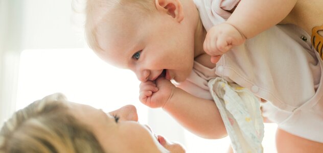 Młoda mama spędza konstruktywny czas ze swoim niedawno narodzonym bobasem. Niemowlę bardzo dobrze się bawi wraz z rodzicielką. Zarówno kobieta jak i  noworodek szeroko się uśmiechają.