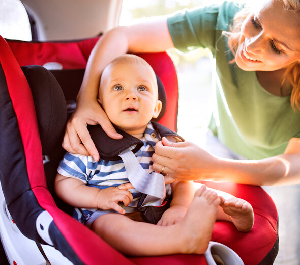 Niemowlę, które ma już kilka miesięcy przygląda się uważnie czemuś interesującemu na podsufitce samochodu. Mama z szerokim uśmiechem starannie zapina 5 punktowe pasy niemowlaka.