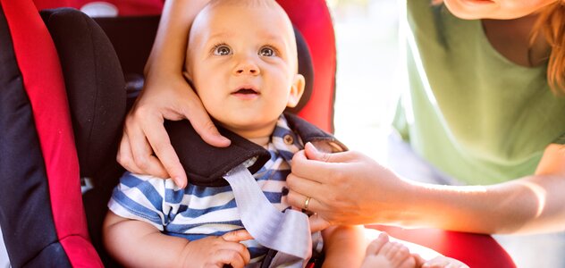 Niemowlę, które ma już kilka miesięcy przygląda się uważnie czemuś interesującemu na podsufitce samochodu. Mama z szerokim uśmiechem starannie zapina 5 punktowe pasy niemowlaka.