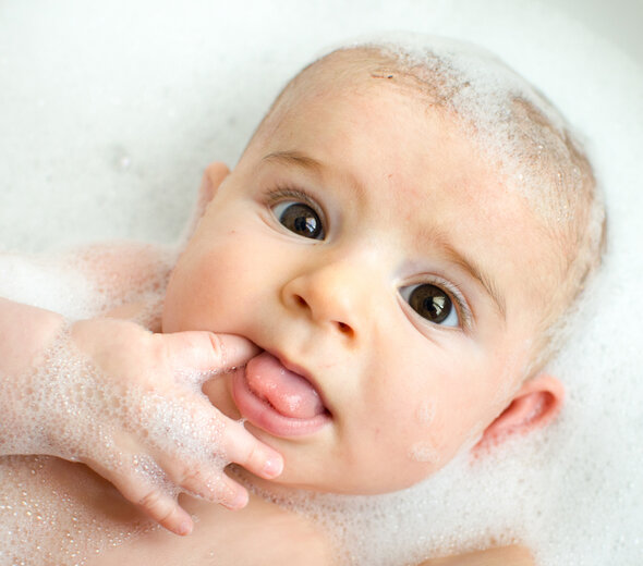 Brązowookie niemowlę leży w wanience pełnej wody i otoczone jest dużą ilością piany. Dziecko trzyma paluszek w buzi i patrzy szeroko otwartymi oczami prosto w obiektyw.