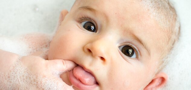 Brązowookie niemowlę leży w wanience pełnej wody i otoczone jest dużą ilością piany. Dziecko trzyma paluszek w buzi i patrzy szeroko otwartymi oczami prosto w obiektyw.