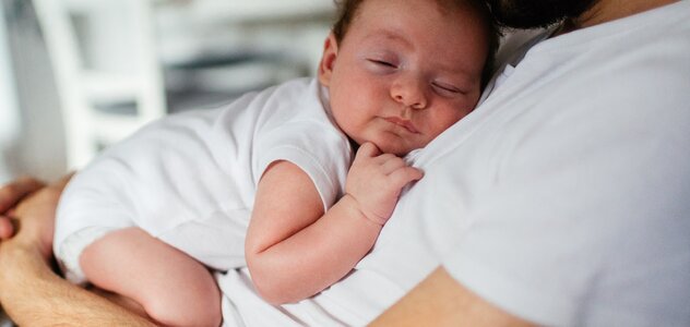 Dziecko spokojnie usypia na brzuchu ojca, który przytula je czule. Niemowlę jest ubrane w białe body i trzyma rączkę blisko buzi. Mężczyzna całuje główkę malucha.