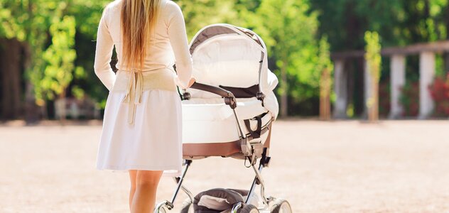 Matka jest na spacerze w parku wraz z dzieckiem, które znajduje się w biało-beżowym wózku. Kobieta jest ubrana w lekką, białą sukienkę oraz jasny sweterek.