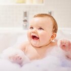 Niemowlę siedzi w wannie z wodą, pełną piany. Dziecko trzyma w powietrzu rączki otoczone gęstą pianą oraz uśmiecha się, przez co wygląda na szczęśliwe i spokojne.