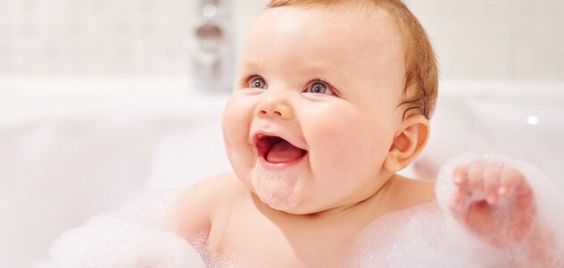 Niemowlę siedzi w wannie z wodą, pełną piany. Dziecko trzyma w powietrzu rączki otoczone gęstą pianą oraz uśmiecha się, przez co wygląda na szczęśliwe i spokojne.