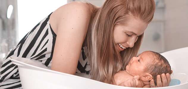 Matka trzyma delikatnie niemowlę w rękach i zanurza je w wanience z wodą, aby je wykąpać. Kobieta uśmiecha się do maluszka, który rozgląda się z zaciekawieniem.