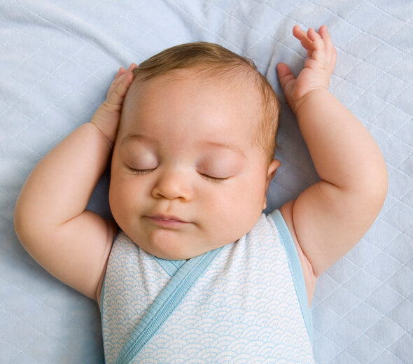 Niemowlę śpi na łóżku, ubrane w niebieskie body. Dziecko trzyma rączki w górze, dotykając swojej główki. Spokojny sen jest dla maluszka bardzo ważny w procesie rozwoju.