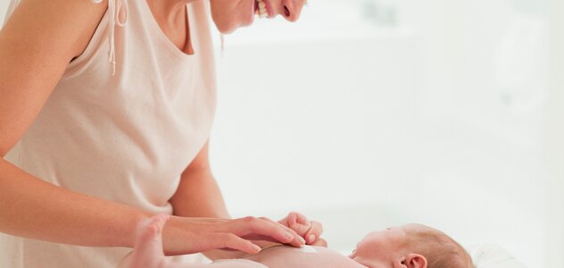 Młoda, uśmiechnięta kobieta nachyla się nad leżącym w pieluszce niemowlęciem. Matka delikatnie smaruje brzuszek dziecka kremem. Nawilżenie jest bardzo ważne dla skóry.