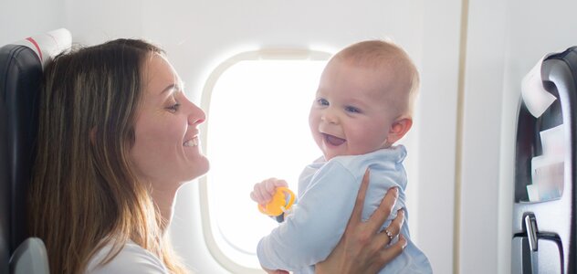 Młoda, uśmiechnięta kobieta siedzi w samolocie wraz z dzieckiem. Oboje uśmiechają się, podczas gdy matka zabawia dziecko, aby nie stresowało się nowym doświadczeniem.