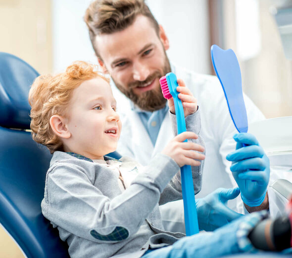 Kilkuletni chłopczyk siedzi na fotelu dentystycznym w gabinecie stomatologicznym. Dziecko przygląda się uważnie dużej, sztucznej szczotce do mycia zębów, obok chłopca siedzi dentysta.