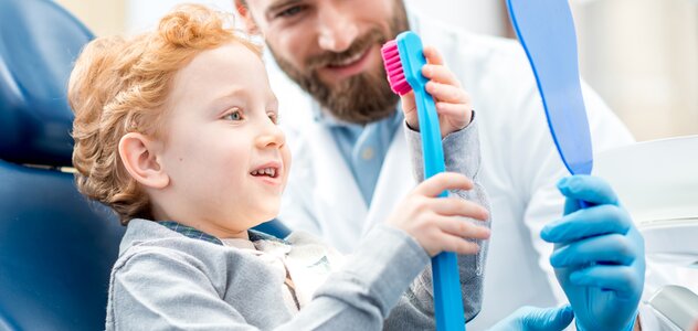 Kilkuletni chłopczyk siedzi na fotelu dentystycznym w gabinecie stomatologicznym. Dziecko przygląda się uważnie dużej, sztucznej szczotce do mycia zębów, obok chłopca siedzi dentysta.