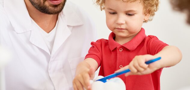 kilkunastomiesięczny chłopiec przebywa w gabinecie stomatologicznym. Młodzieniec uczy się poprawnie dbać o swoje zęby. Dziecko dużą szczoteczką myje gigantyczny, sztuczny ząb.