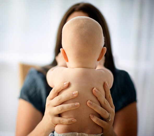 Mama trzyma pod pachami niedawno narodzone niemowlę. Kobieta na wyciągniętych przed siebie rękach trzyma nagiego, ubranego w pieluszkę noworodka. Dziecko wyciąga do mamy ręce.