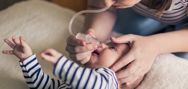 Niedawno narodzone niemowlę leży po środku dużego łóżka rodziców. Noworodek leży na pleckach i stara się przekręcić na bok uciekając od nieprzyjemnej dla niego czynności wyciągania śluzu z nosa.