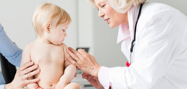 Niedawno narodzony chłopiec cierpliwie i spokojnie siedzi w gabinecie lekarskim. Nieświadomy noworodek patrzy na panią doktor, która stara się ukłuć igłą maluszka w ramię.