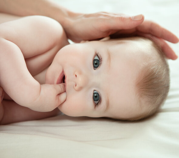 Niedawno narodzony niemowlak leży spokojnie na boczku. Dziecko patrzy na wprost siebie dużymi, szaro-brązowymi oczami i wkłada paluszki do buzi. Mama głaszcze noworodka po główce.