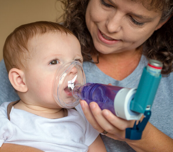 Mama trzyma kilkumiesięcznego malucha na kolanach. Bobas samodzielnie siedzi opierając się o rodzicielkę. Kobieta przystawia bobasowi do buzi inhalator z podłączonym do niego lekarstwem na astmę.