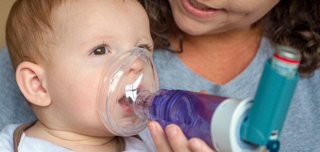 Mama trzyma kilkumiesięcznego malucha na kolanach. Bobas samodzielnie siedzi opierając się o rodzicielkę. Kobieta przystawia bobasowi do buzi inhalator z podłączonym do niego lekarstwem na astmę.