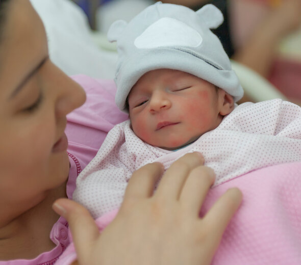 Niedawno narodzone niemowlę leży na rękach u mamy. Noworodek zawinięty jest starannie w jasny kocyk i ma założoną czapeczkę. Mama przytula do swojej piersi maluszka.