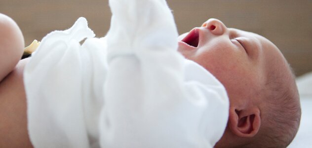 Niedawno narodzone niemowlę leży spokojnie na pleckach na dużej macie do badania niemowląt. Noworodek ubrany jest w kaftanik na długi rękaw. Pępek bobasa jeszcze samoistnie nie odpadł.