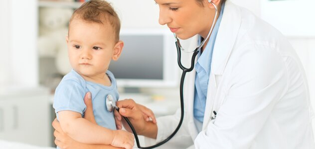 Niedawno narodzony niemowlak samodzielnie siedzie na kozetce w gabinecie lekarskim. pani doktor, która stoi na przeciw malucha podtrzymuje go pod pachami i bada stetoskopem.