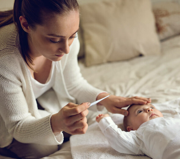 Młoda mama siedzi na dużym, małżeńskim łożu. Obok kobiety po środku materaca leży na pleckach niedawno narodzone niemowlę. Rodzicielka trzyma jedną rękę na czole malucha a w drugiej trzyma termometr.