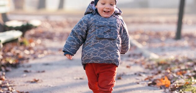 Kilkumiesięczny chłopiec spaceruje po parku. Dziecko raźnym krokiem z szerokim uśmiechem na twarzy przechadza się po ścieżkach pomiędzy drzewami w okresie jesiennym.