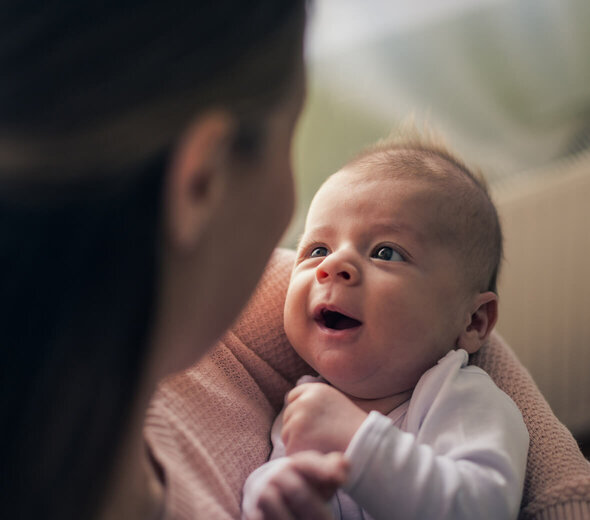 Młoda mama przebywa z niedawno narodzonym niemowlakiem w salonie swojego domu. Kobieta siedzi na sofie i trzyma na rękach malucha. Noworodek się serdecznie uśmiecha do rodzicielki. 