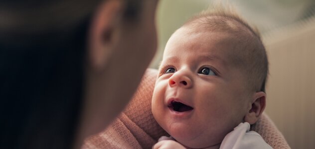 Młoda mama przebywa z niedawno narodzonym niemowlakiem w salonie swojego domu. Kobieta siedzi na sofie i trzyma na rękach malucha. Noworodek się serdecznie uśmiecha do rodzicielki. 