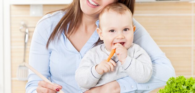 Młoda mama zajmuje się gotowaniem obiadu dla niedawno narodzonego maluszka. Noworodek przebywa w kuchni razem z rodzicielką. Kobieta stoi za blatem, na blacie siedzi bobas a dookoła rozłożone jest jedzenie.