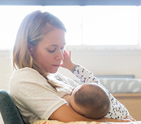 Młoda mama przebywa w salonie trzymając na rękach niedawno narodzone niemowlę. Maluszek spokojnie leży na rękach rodzicielki i wyciąga w stronę kobiety malutką rączkę.