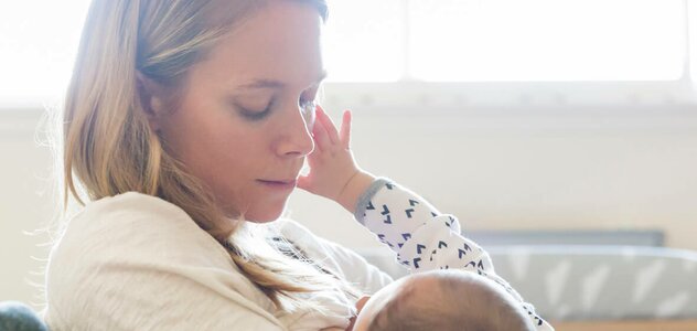 Młoda mama przebywa w salonie trzymając na rękach niedawno narodzone niemowlę. Maluszek spokojnie leży na rękach rodzicielki i wyciąga w stronę kobiety malutką rączkę.