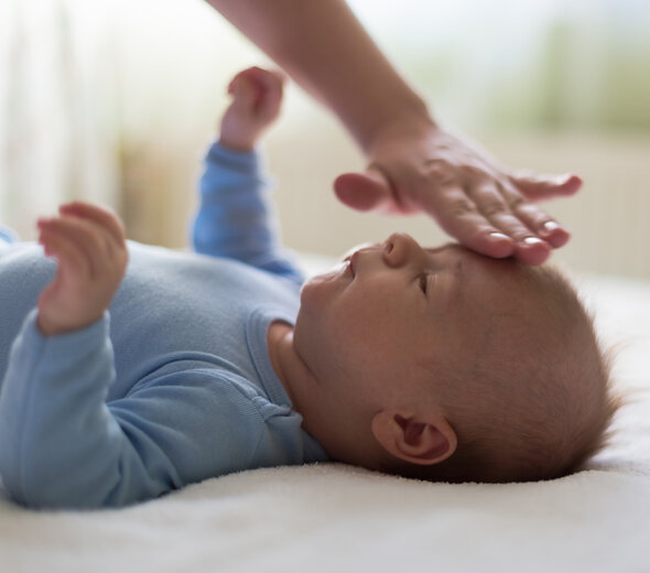 Niedawno narodzony maluch leży po środku dużego, miękkiego, dwuosobowego łóżka rodziców. Mama przykłada dziecku rękę do czoła w celu sprawdzenia stanu jego zdrowia. 