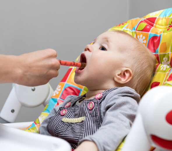 Niedawno narodzony maluch samodzielnie siedzi w kuchni. Dziecko siedzi w krzesełku do karmienia wyściełanym pokrowcem. Maluch otwiera szeroko buzię żeby przyjąć dawkę lekarstwa.