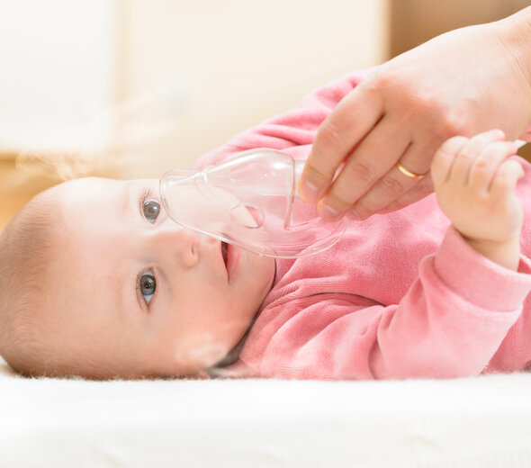 Młoda mama podstawia swojemu niedawno narodzonemu niemowlakowi ustnik do inhalacji dzieci do buzi. Noworodek z uśmiechem na twarzy przyjmuje przynoszącą ulgę inhalację.