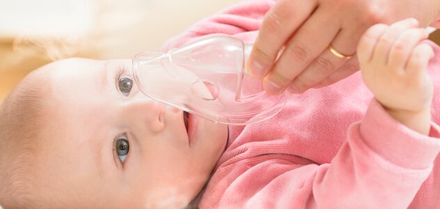 Młoda mama podstawia swojemu niedawno narodzonemu niemowlakowi ustnik do inhalacji dzieci do buzi. Noworodek z uśmiechem na twarzy przyjmuje przynoszącą ulgę inhalację.