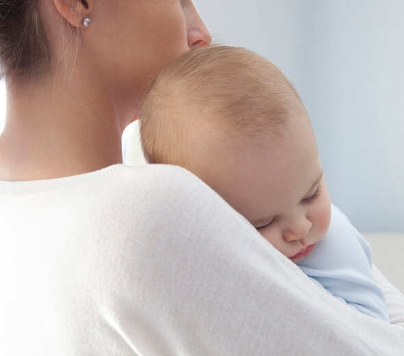 Młoda matka delikatnie przytula do siebie spokojnie śpiące niemowlę. Więź matki z dzieckiem umożliwia maluszkowi zachowanie spokoju w trudnych dla siego sytuacjach.