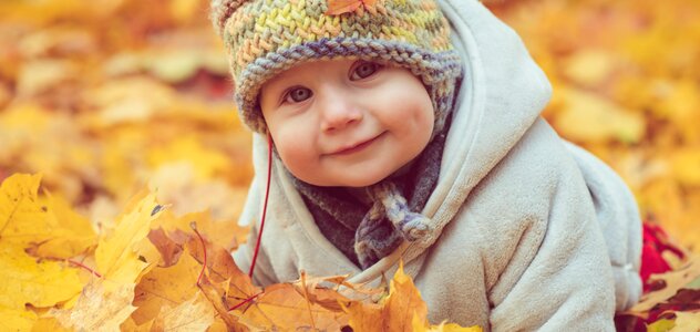 Uśmiechnięte niemowlę siedzi na ziemi w parku wśród sterty pożółkłych liści. Maluszek ma na główce kolorową wełnianą czapkę, na której leży pomarańczowy listek.