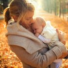 Młoda matka delikatnie przytula do siebie niemowlę, które ma na sobie pajacyka oraz wełniany sweterek. Kobieta wraz z dzieckiem stoją na tle jesiennego krajobrazu.