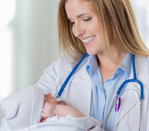 Uśmiechnięta, młoda lekarka trzyma na rękach spokojnie śpiące niemowlę, które owinięte jest w kocyk. Kobieta ma na sobie niebieską koszulę oraz biały fartuch.