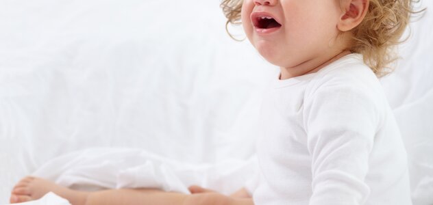 Dziecko siedzi na białej pościeli łóżka w białym body na długi rękaw. Maluszek ma minkę, która wyraża niezadowolenie, które może być wynikiem bólu lub dyskomfortu.